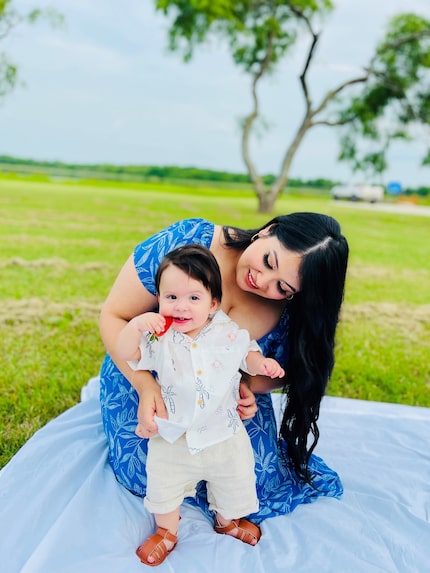 A young mother in a blue dress kneels on a blanket spread on the grass and supports her...