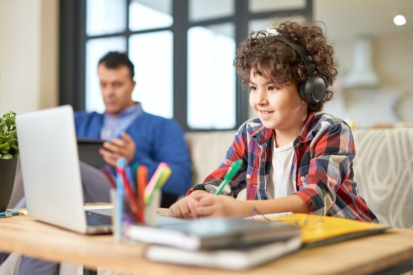 Enthusiastic Latin American boy wearing headphones and smiling while doing a school task on...