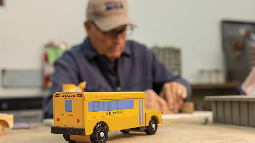 Man handcrafts a wooden gift while a completed bus toy sits in the foreground