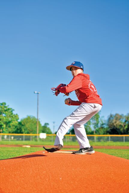 12-year-old Austin prepares to throw a baseball on a baseball field, dressed in his red...
