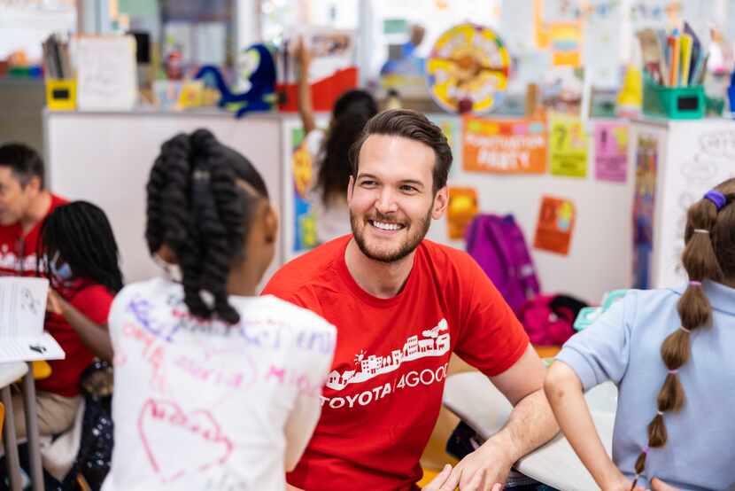 Toyota volunteer in red T-shirt sits at a classroom table talking with a female elementary...
