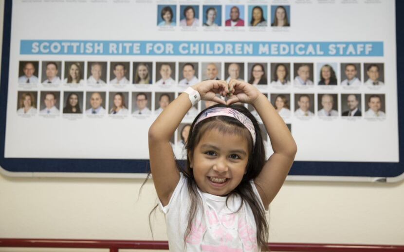 With her hands positioned into a heart, a young girl poses in front of a collage of photos...