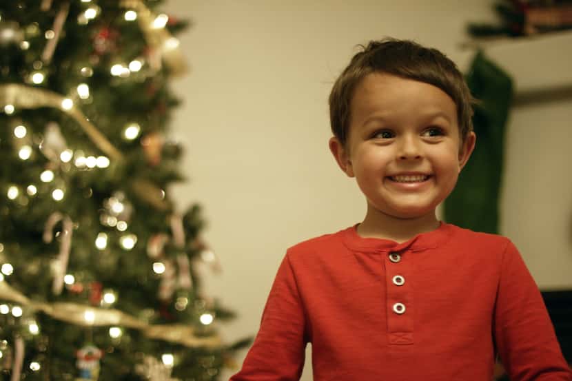 A 5-year-old boy in a red shirt smiles in front of a Christmas tree.