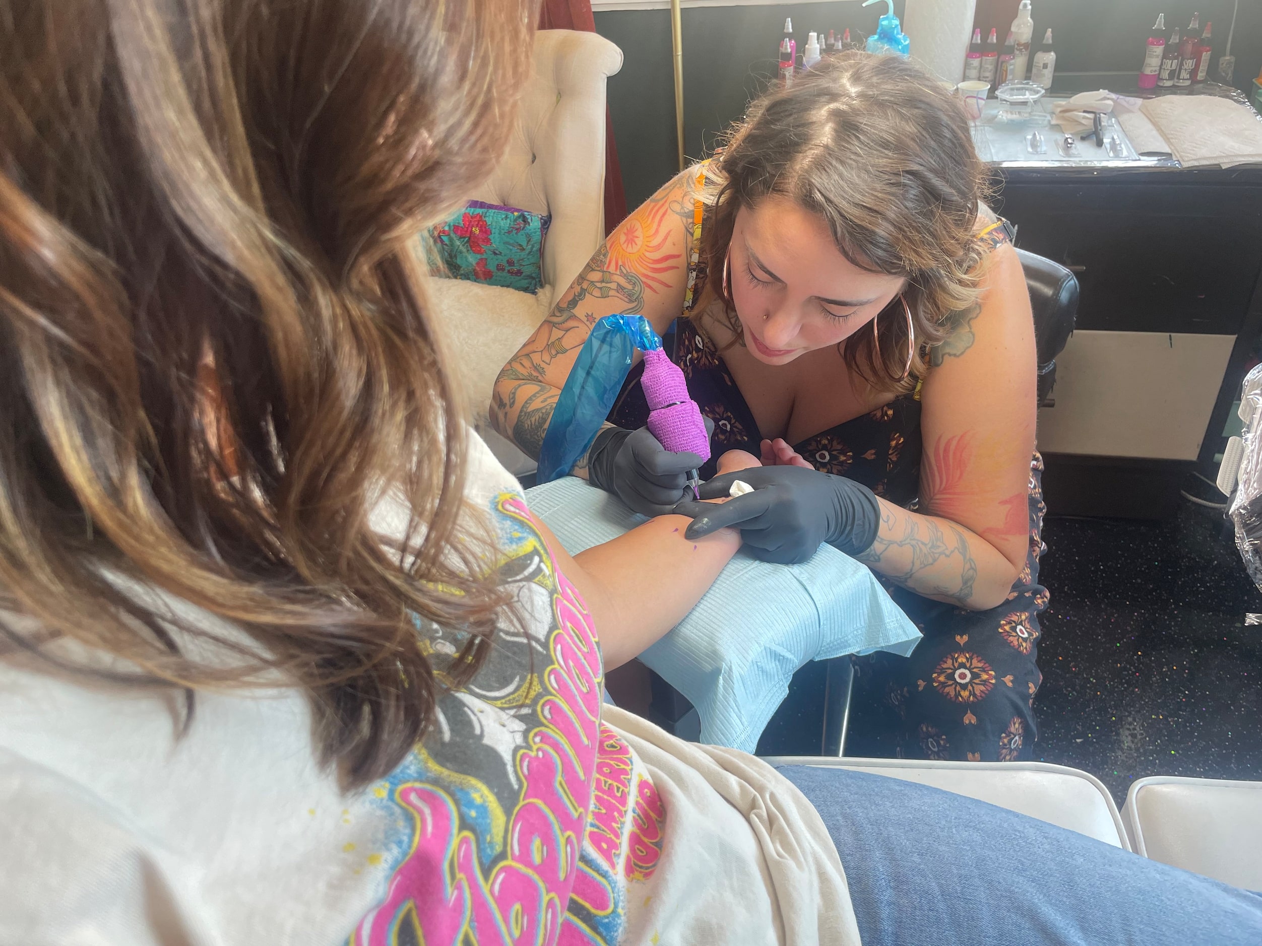 Woman sits in tattoo artist's chair as the artist leans over her applying a tattoo to her...