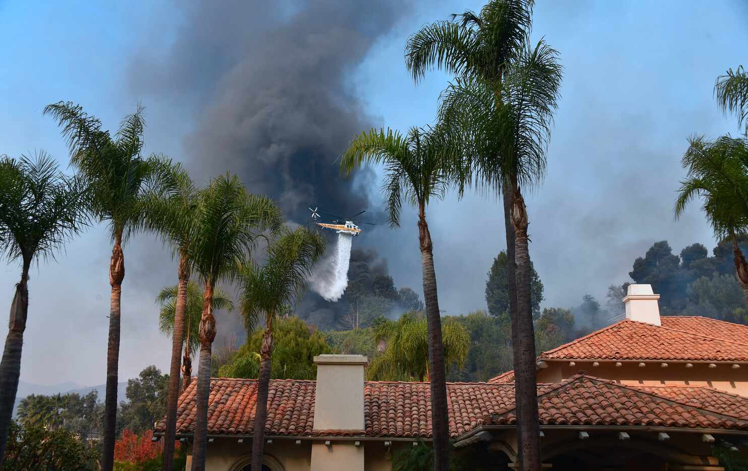 A helicopter drops water over burning homes in Bel Air as a huge plume of black smoke rises...