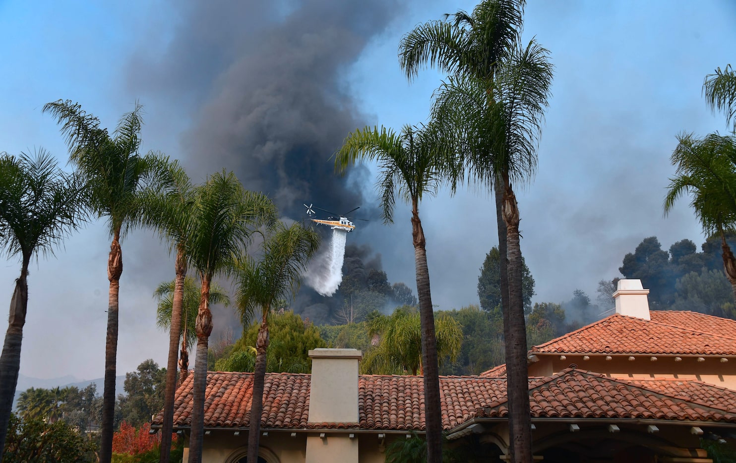 A helicopter drops water over burning homes in Bel Air as a huge plume of black smoke rises...