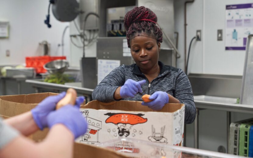 A worker in a hair net wearing purple gloves peels vegetables in a kitchen as part of...