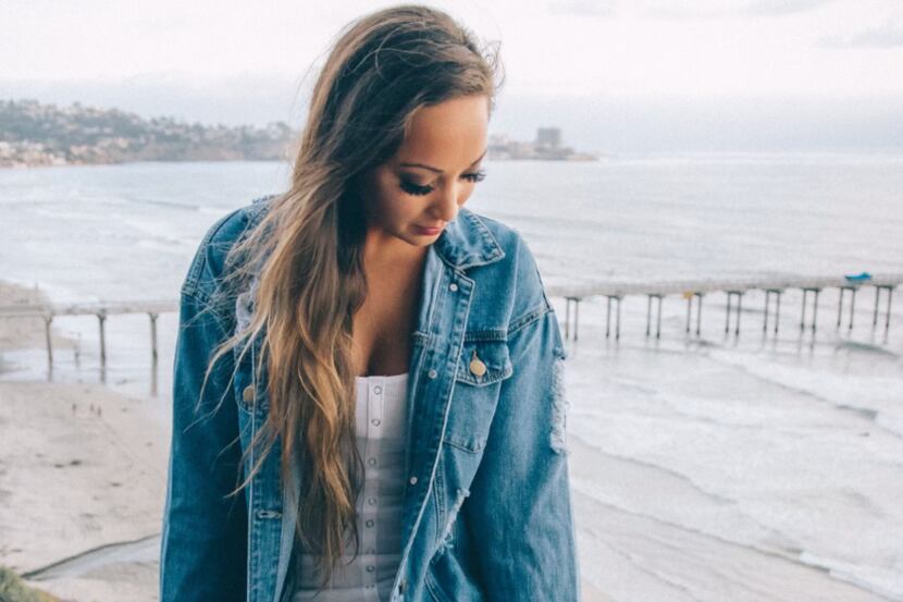 Sarah P Antonella wears a denim jacket while looking down while the beach is in the background