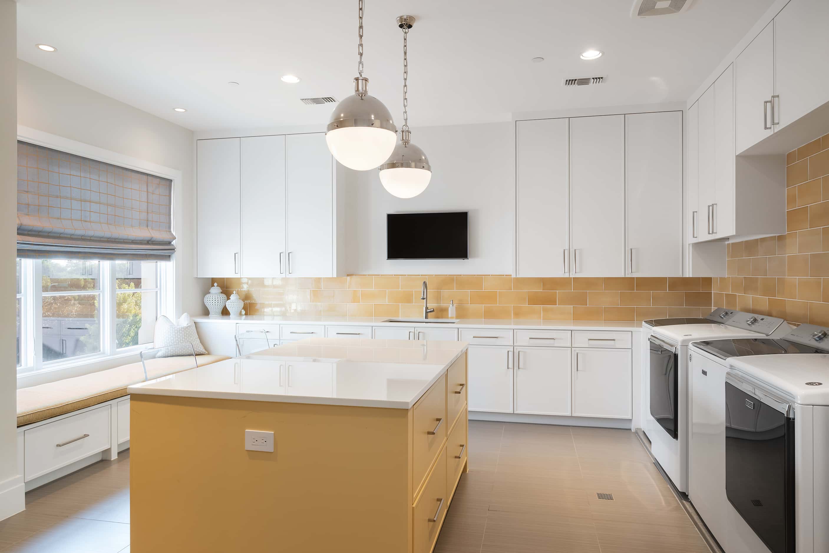Laundry room with white appliances, white cabinets, a yellow backsplash and a yellow island.