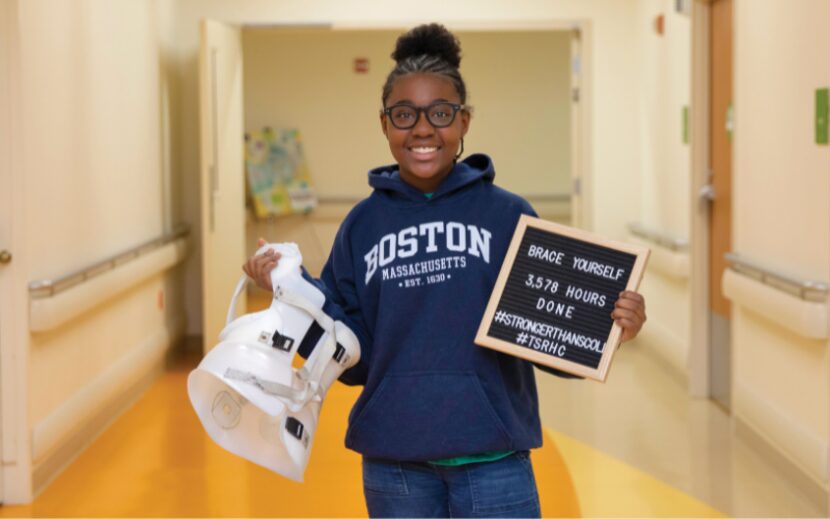 A young girl holds a scoliosis brace and a sign that reads "Brace Yourself, 3.578 hours...