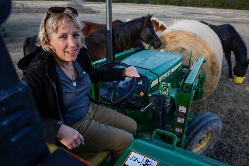 Brenda Rizos poses while moving a bale of hay around with a tractor surrounded by horses on...
