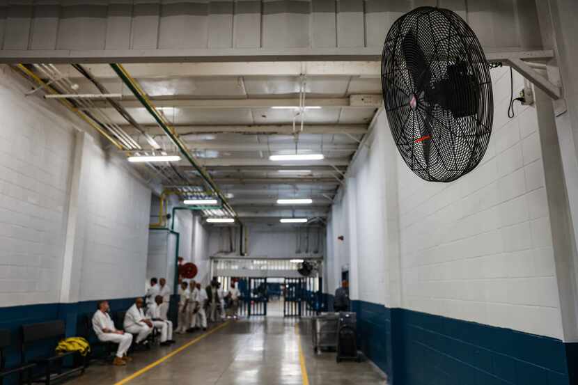 Inmates dressed in white sit along a bench on the left side of a long hallway ending in...