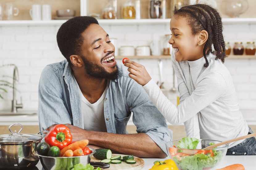 Young girl feeding dad in kitchen, giving him cherry tomato while making salad.