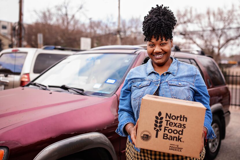 A woman holds a cardboard box that reads North Texas Food Bank in front of a maroon car.