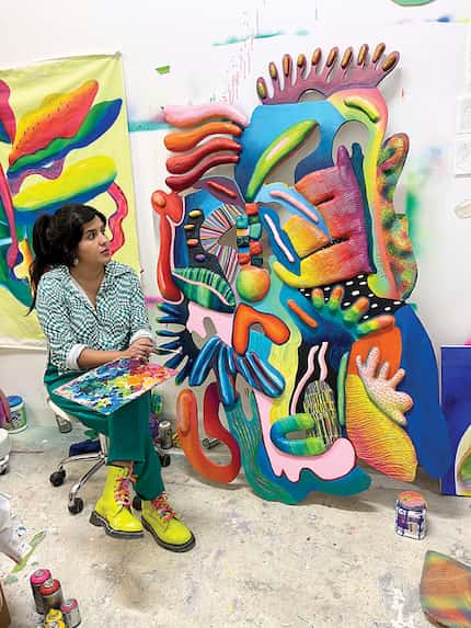 Woman dressed in green with bright yellow boots sits next to a colorful mixed-media artwork.