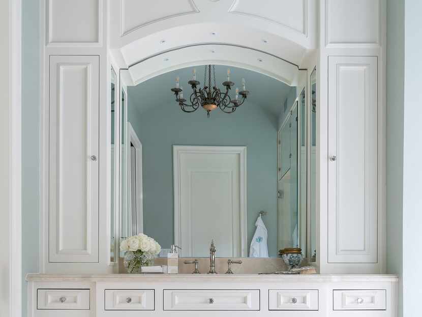 White bathroom cabinets with pale blue wall reflected in a mirror, with black chandelier...