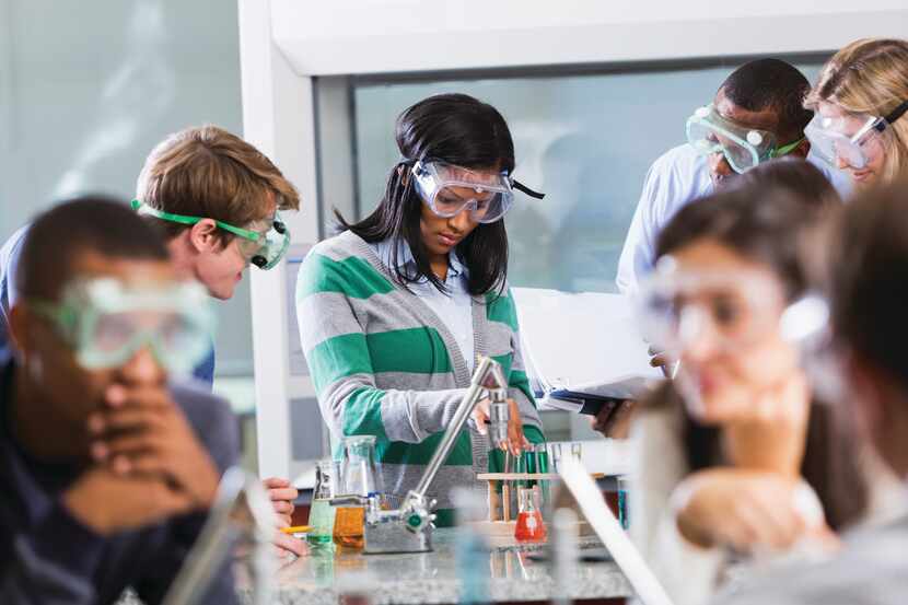 Multi-ethnic group of students with instructor in chemistry lab, wearing safety goggles,...