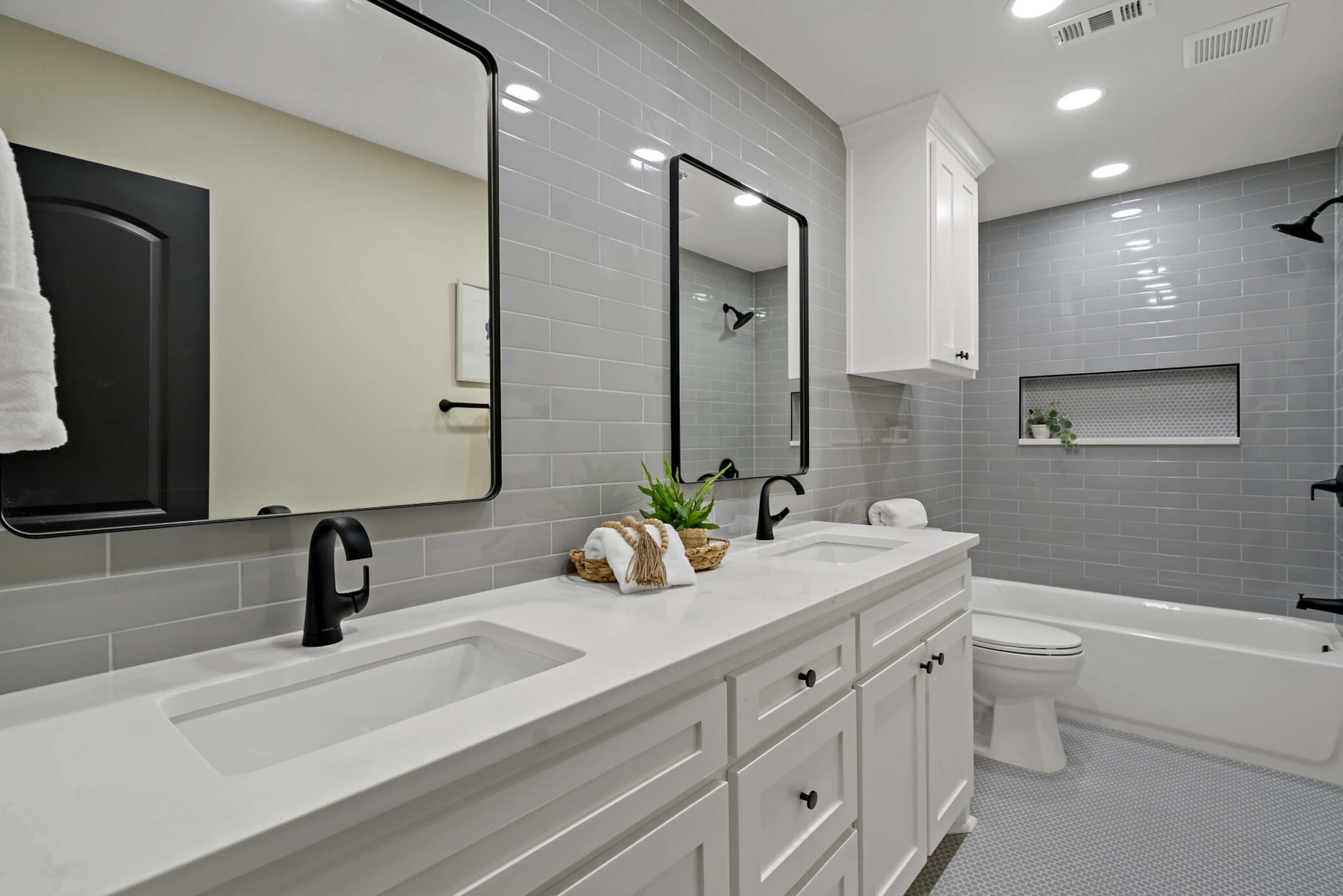 Bathroom with gray tile on walls and floors, white countertops and cabinetry, dual sinks,...