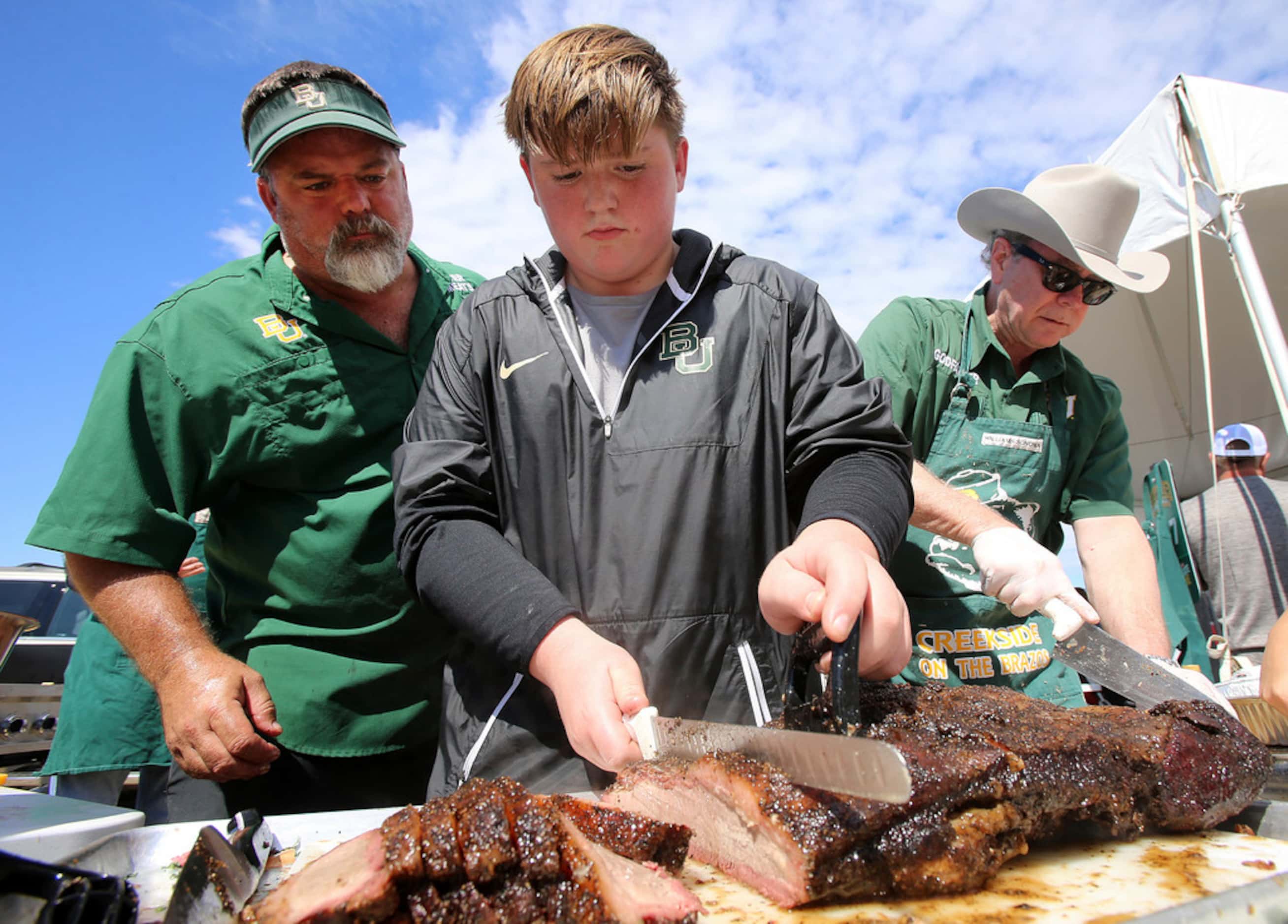 Luke Grant, 14, slices up brisket as his father Jonathan, left, looks on at their tailgate...