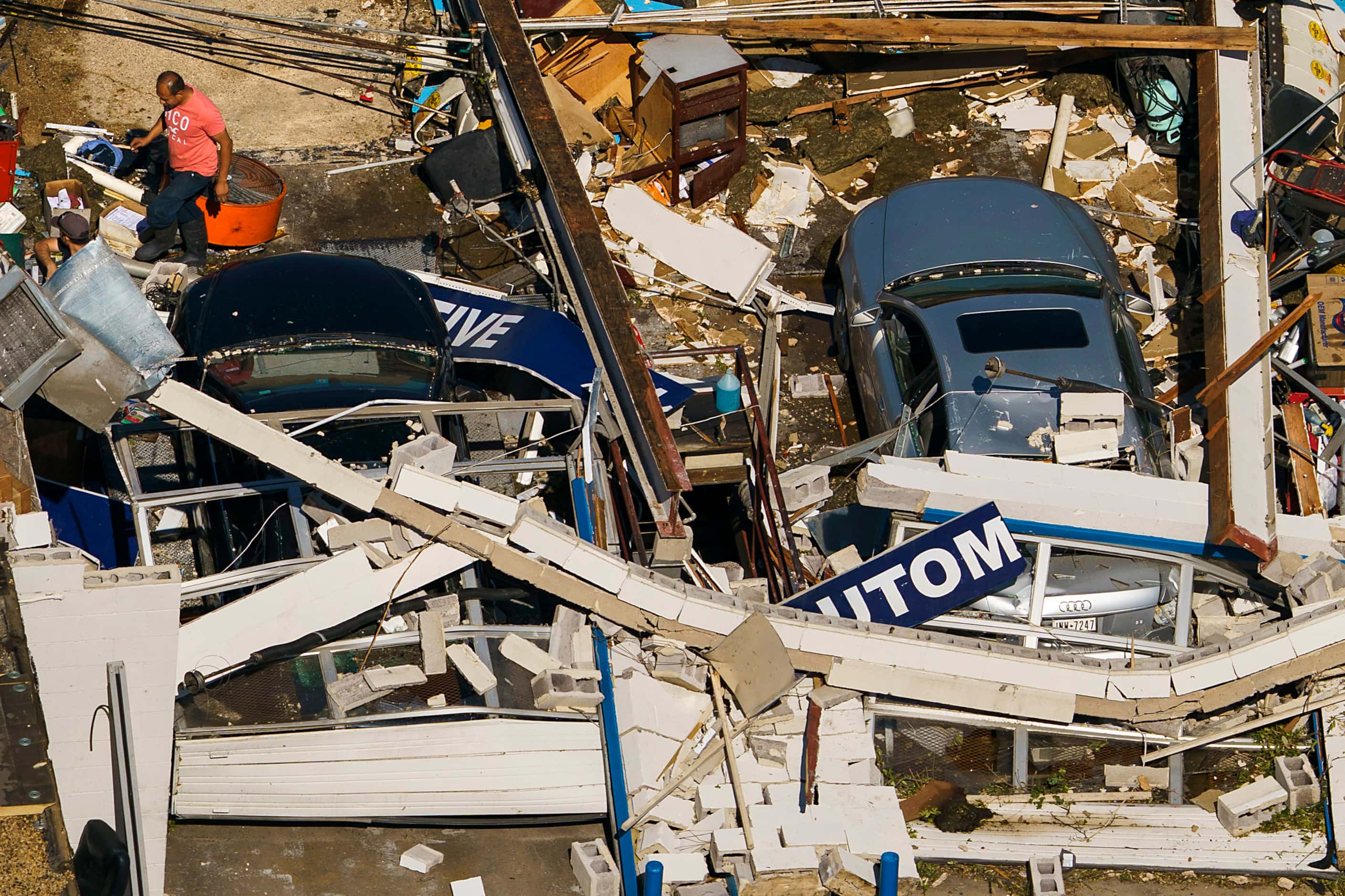 People walk amidst cars buried in the rubble of a destroyed auto repair shop on Northaven...