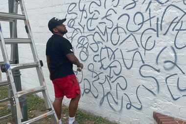 A Fort Worth-based artist stands by a ladder and looks at a white wall. He's holding a spray...