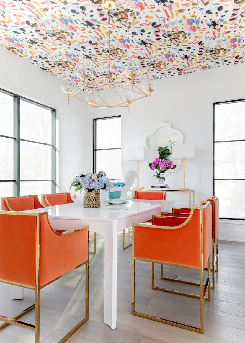 A dining room has orange chairs and a ceiling with multi-colored patterned wallpaper.