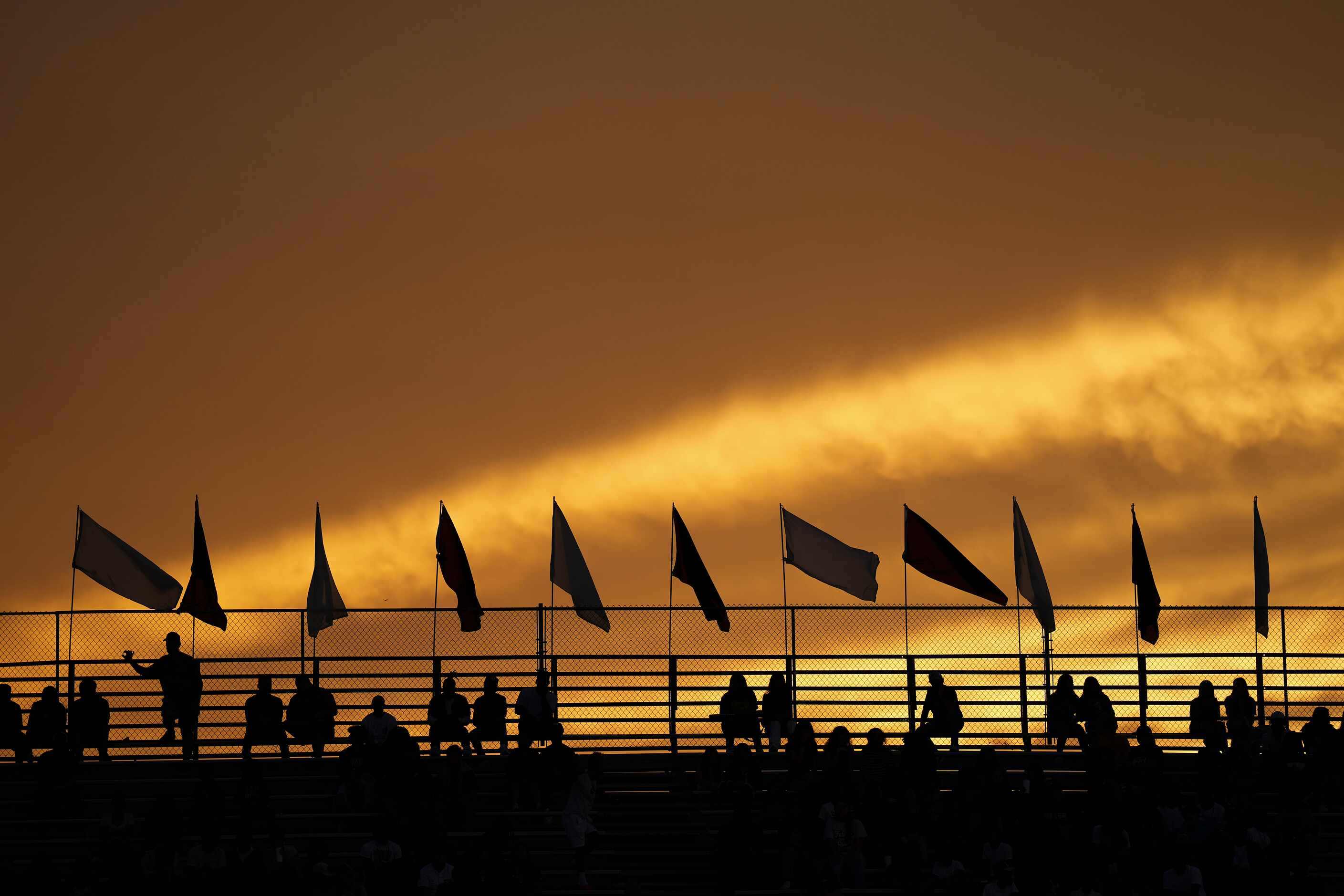 Fans watch from atop the stands as the sun sets on a high school football game between...