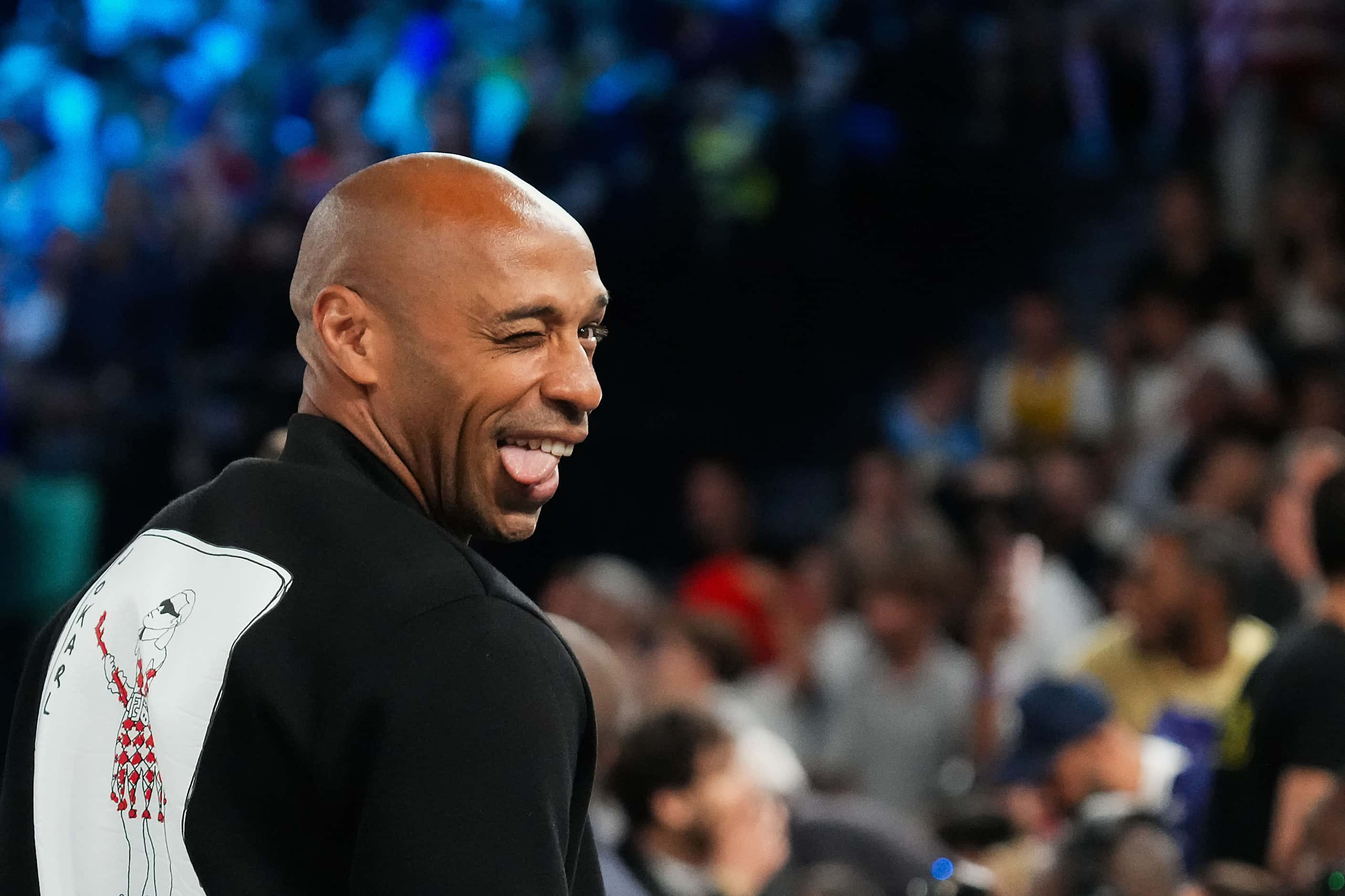 Thierry Henry makes a face toward Dirk Nowitzki as they watch from courtside during the...