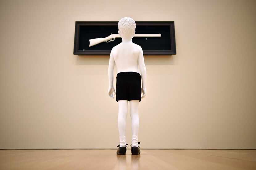 In One Day, a shirtless boy gazes up at a rifle mounted on the wall. The artists say that...