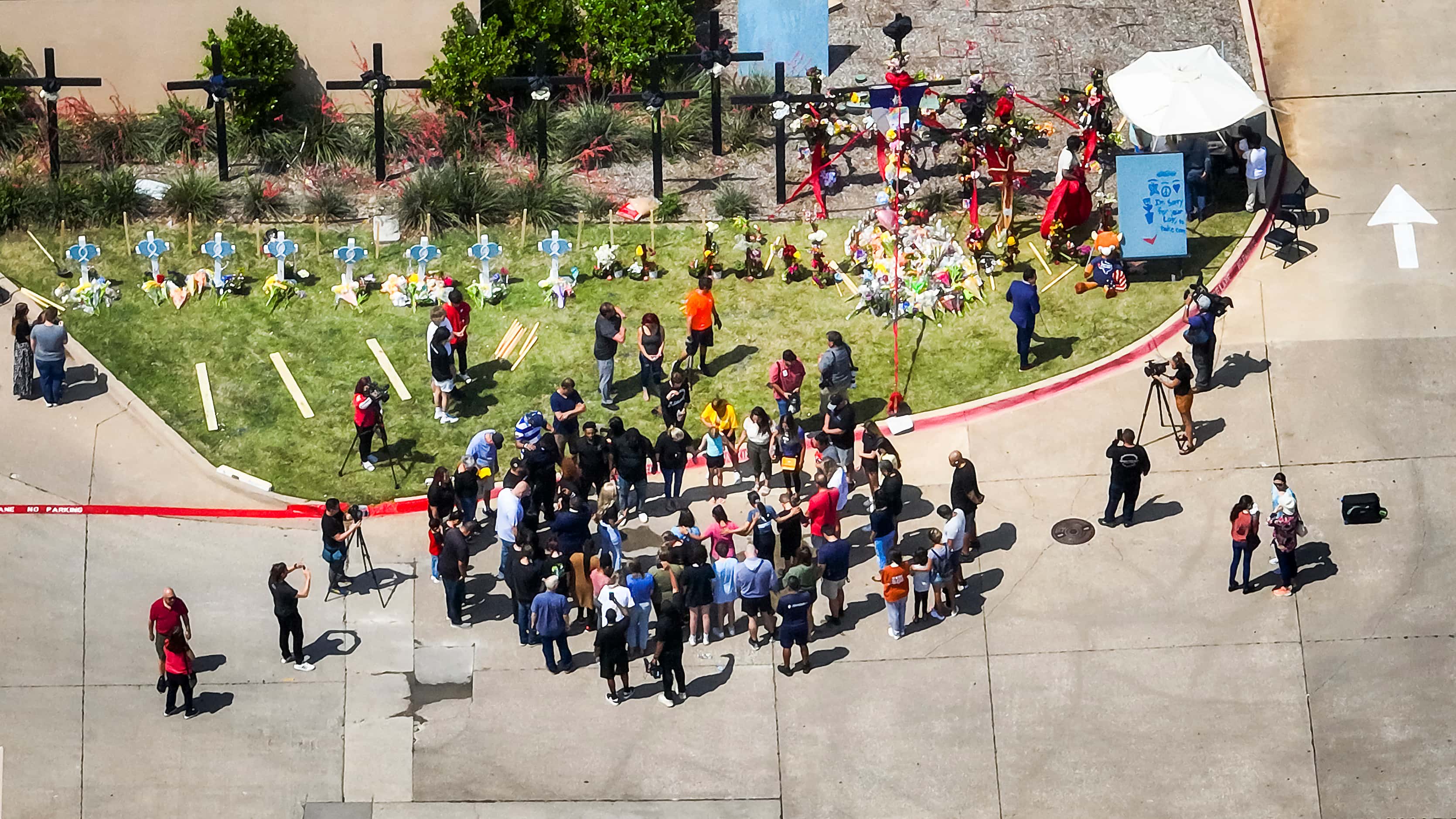 Aerial view of people gathering in prayer near a growing memorial outside the Allen Premium...