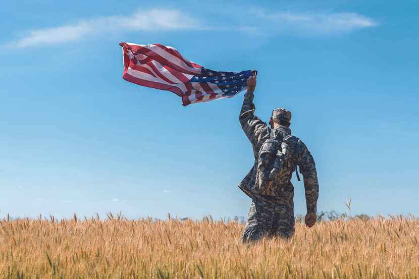 An Army person in uniform walks through a field holding an American flag in the air.