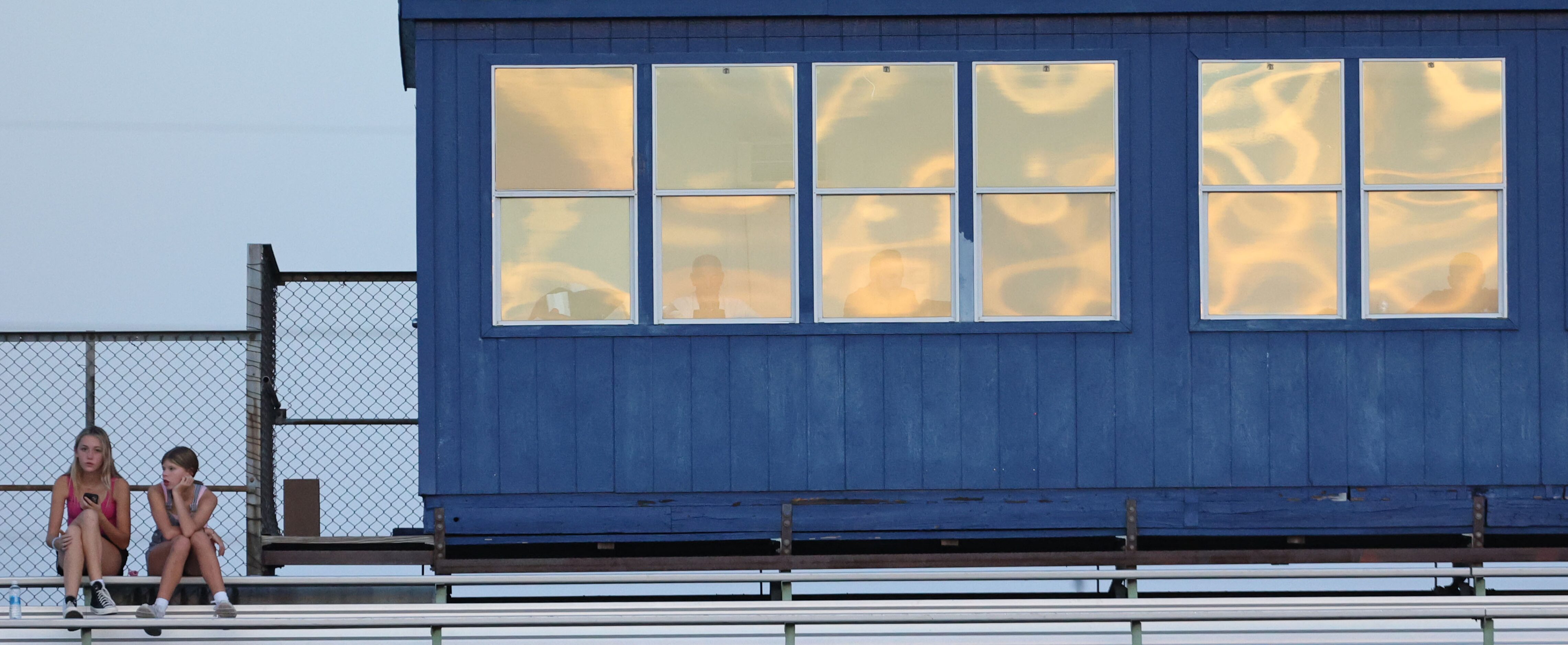 A couple of fans watch game action from the stands, as the sunset is reflected in the...