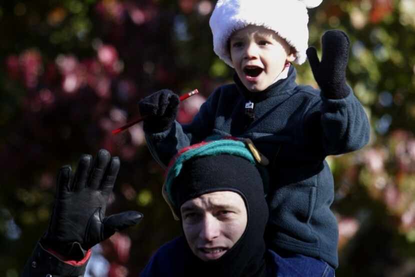 A boy waves while watching a Christmas parade from his father's shoulders.