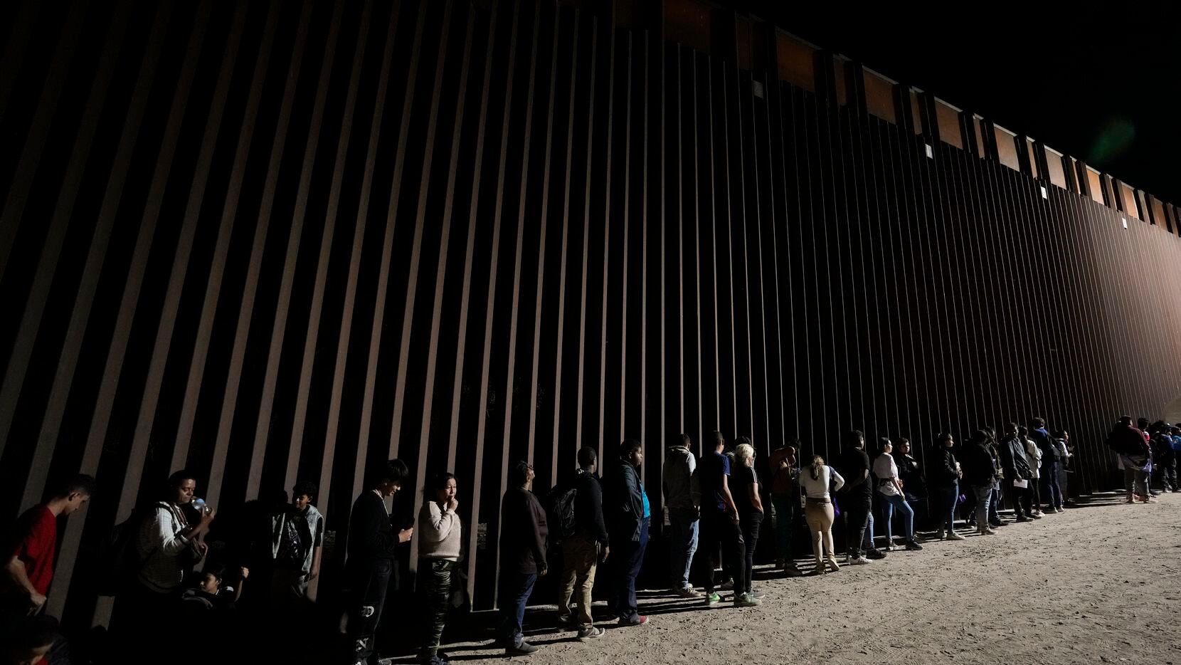Migrantes hacen fila junto al muro fronterizo esperan para solicitar asilo luego de cruzar...