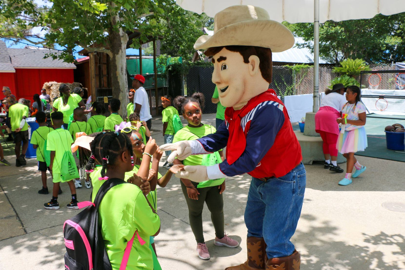 A Big Tex character costume speaks with young children wearing green shirts.